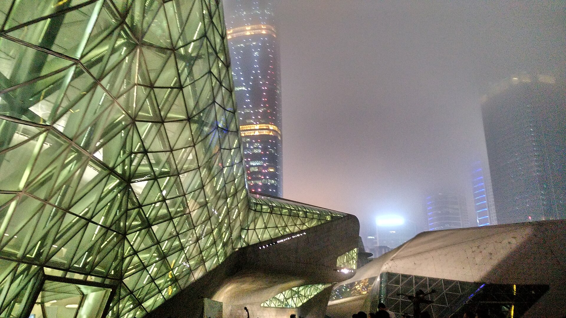 夜雾里，左侧折面的三角网格玻璃幕墙透着内部的钢架与灯光，右侧是灰色混凝土曲面体块，远处两栋摩天楼