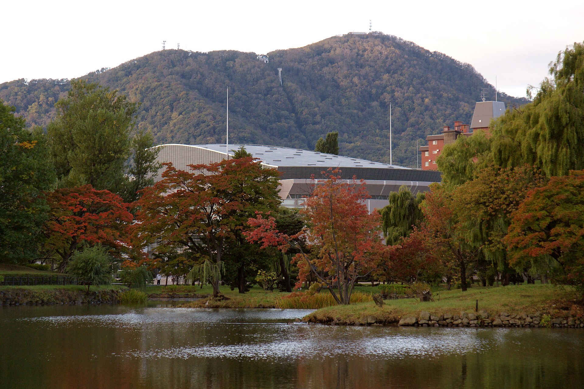 札幌中岛公园的秋日池畔，水面倒映着红黄相间的树叶，弧形金属屋顶的Kitara音乐厅立于林间，远处是带天线的山影。