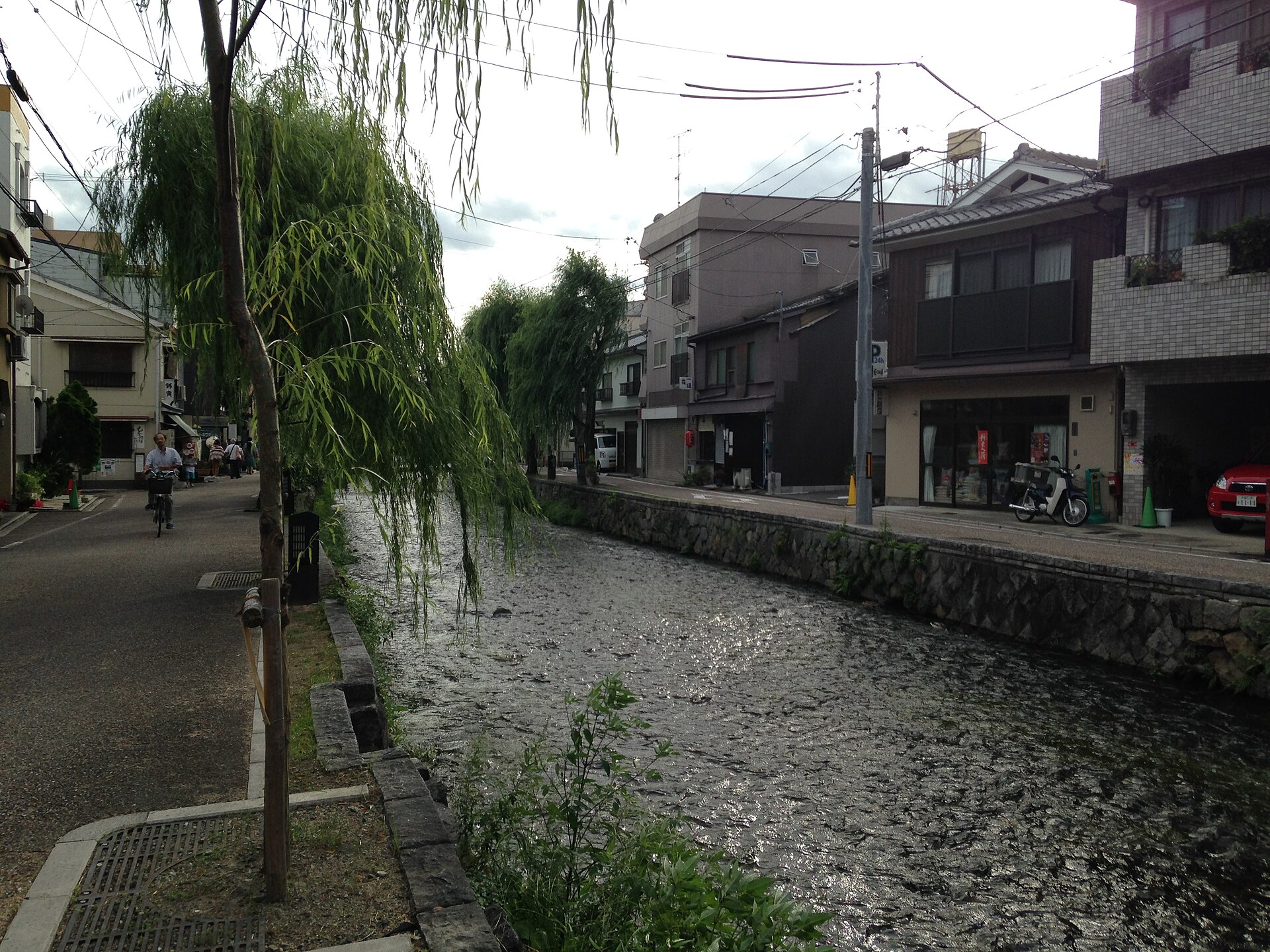 窄水道夹在砌石岸之间，左侧垂柳枝条垂落、有人骑车经过，右侧沿街是深色木格窗的民居与店铺