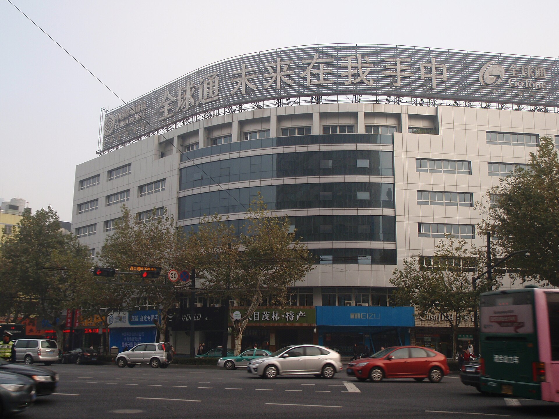 武林路与体育场路交叉口的街景：弧形玻璃幕墙的商业楼、底层沿街店铺与成排行道树，路口有车辆通行。