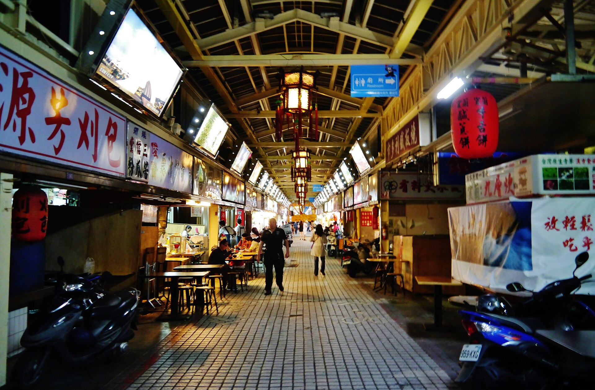 夜市拱廊顶下挂着圆形红灯笼与长条日光灯，两侧饮食摊前摆着不锈钢小方桌与圆凳，招牌可见「源芳刈包」