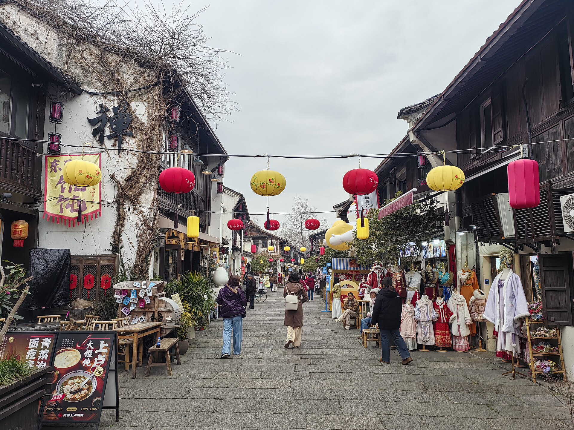 小河直街的青石板窄巷：两侧是白墙黑瓦的传统民居与店铺，檐下悬挂红黄灯笼，巷中有行人往来。
