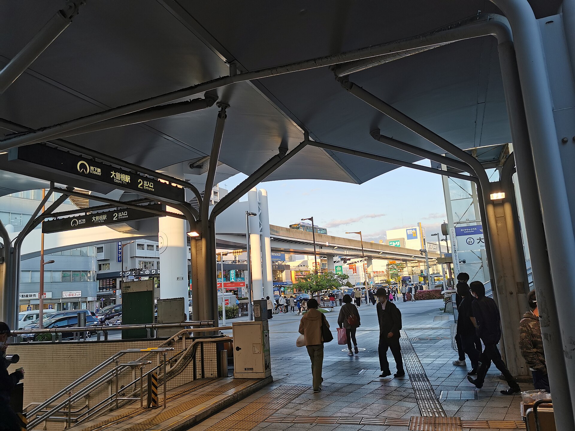 折板天棚下的车站出口，左上方黑底牌子写着「大曽根駅 2 出口」，远处高架梁横过街口