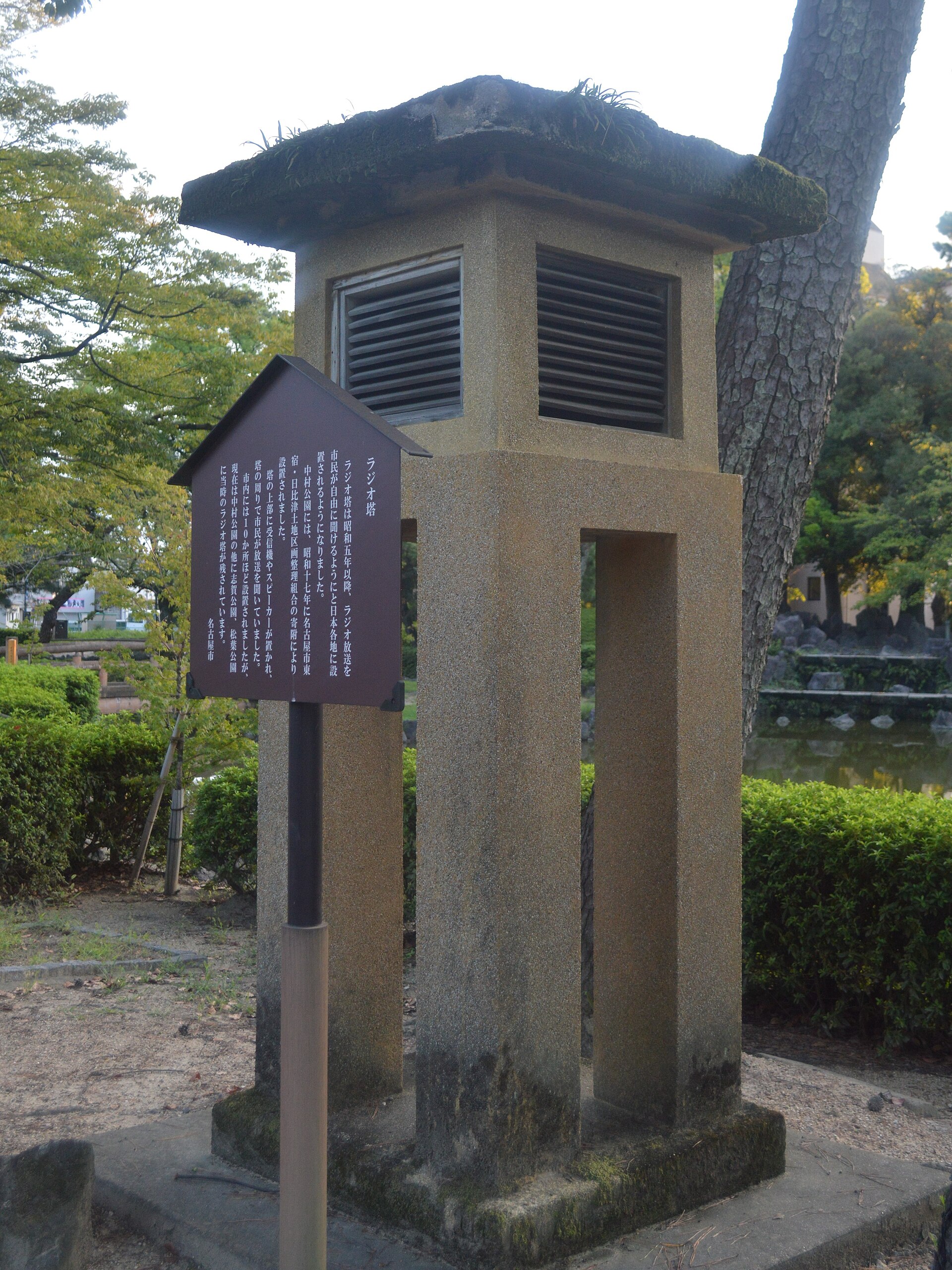 石造方柱塔的下部中空、上部开着百叶口，塔前立着写有「ラジオ塔」「中村公園」的解说牌
