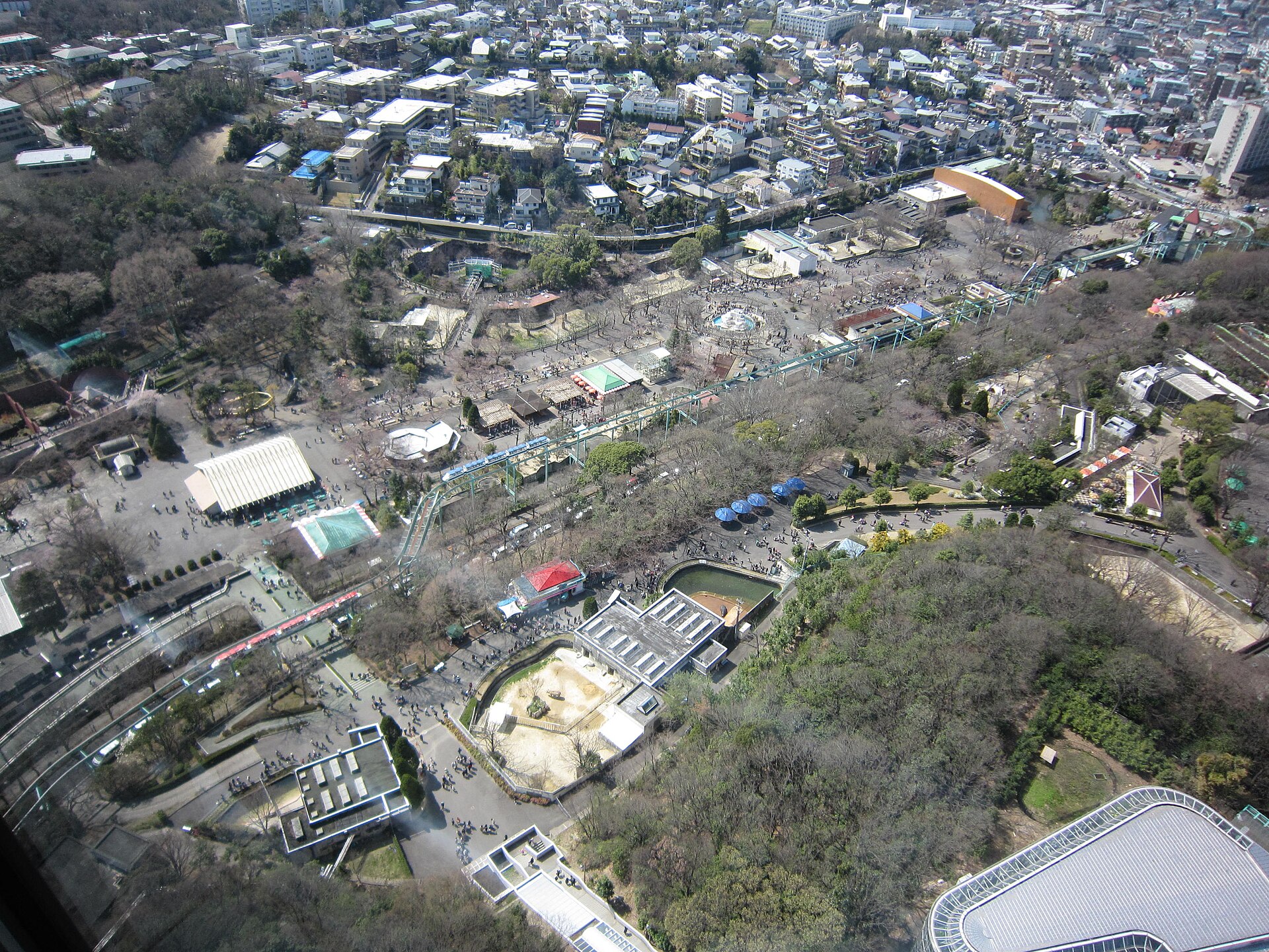 高处俯瞰的公园绿地：树冠大半落尽，园里散着水池、围栏与架空轨道，远处住宅一直铺到天际