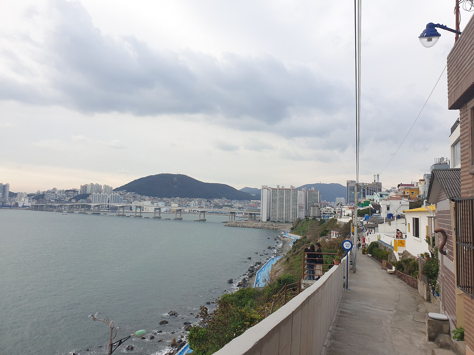 临海坡道从挡墙与铁栏之间往下走，墙外是碎石海岸，远处长桥后立着白色楼群