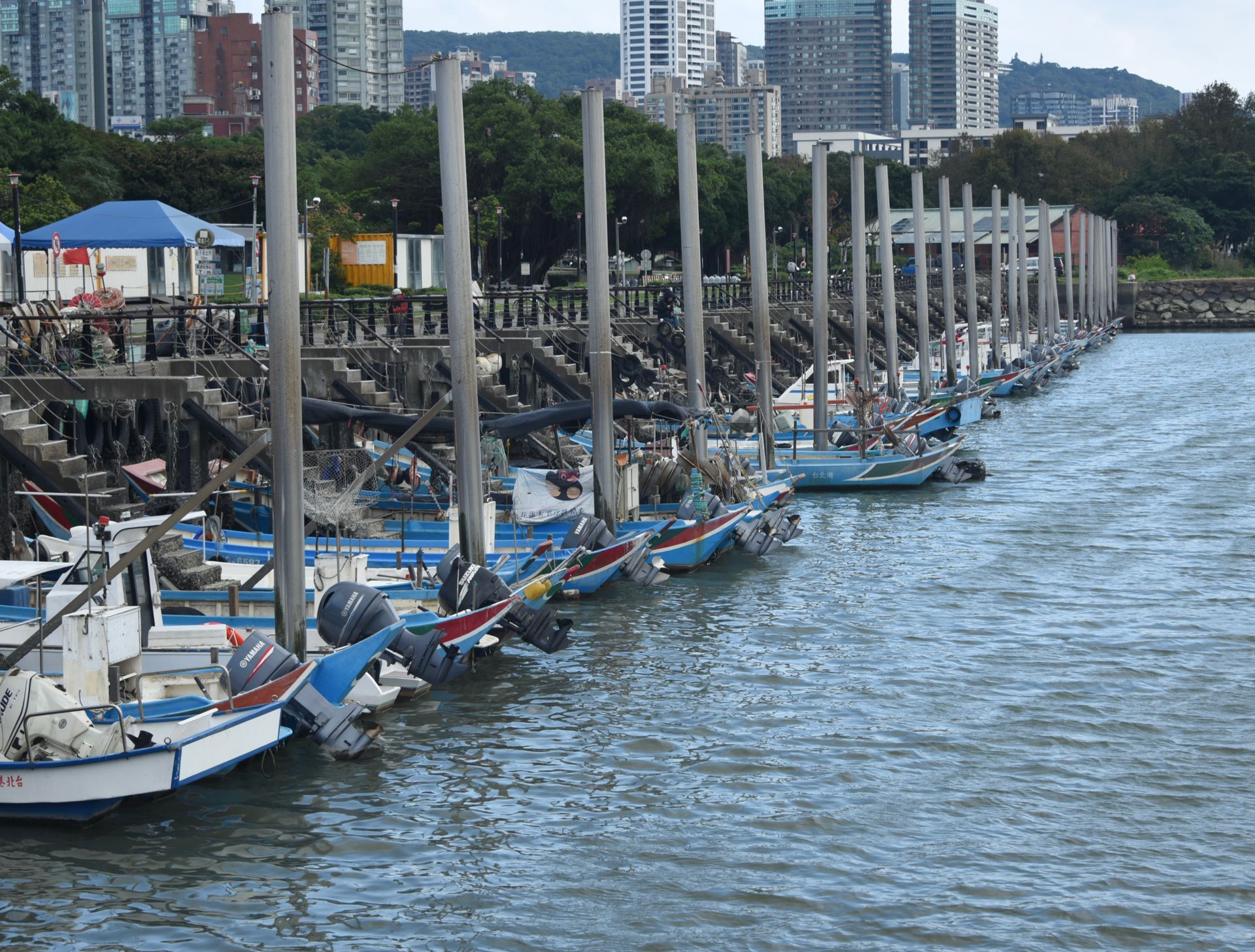 Row of colorful fishing boats docked in an urban marina with city skyline background.