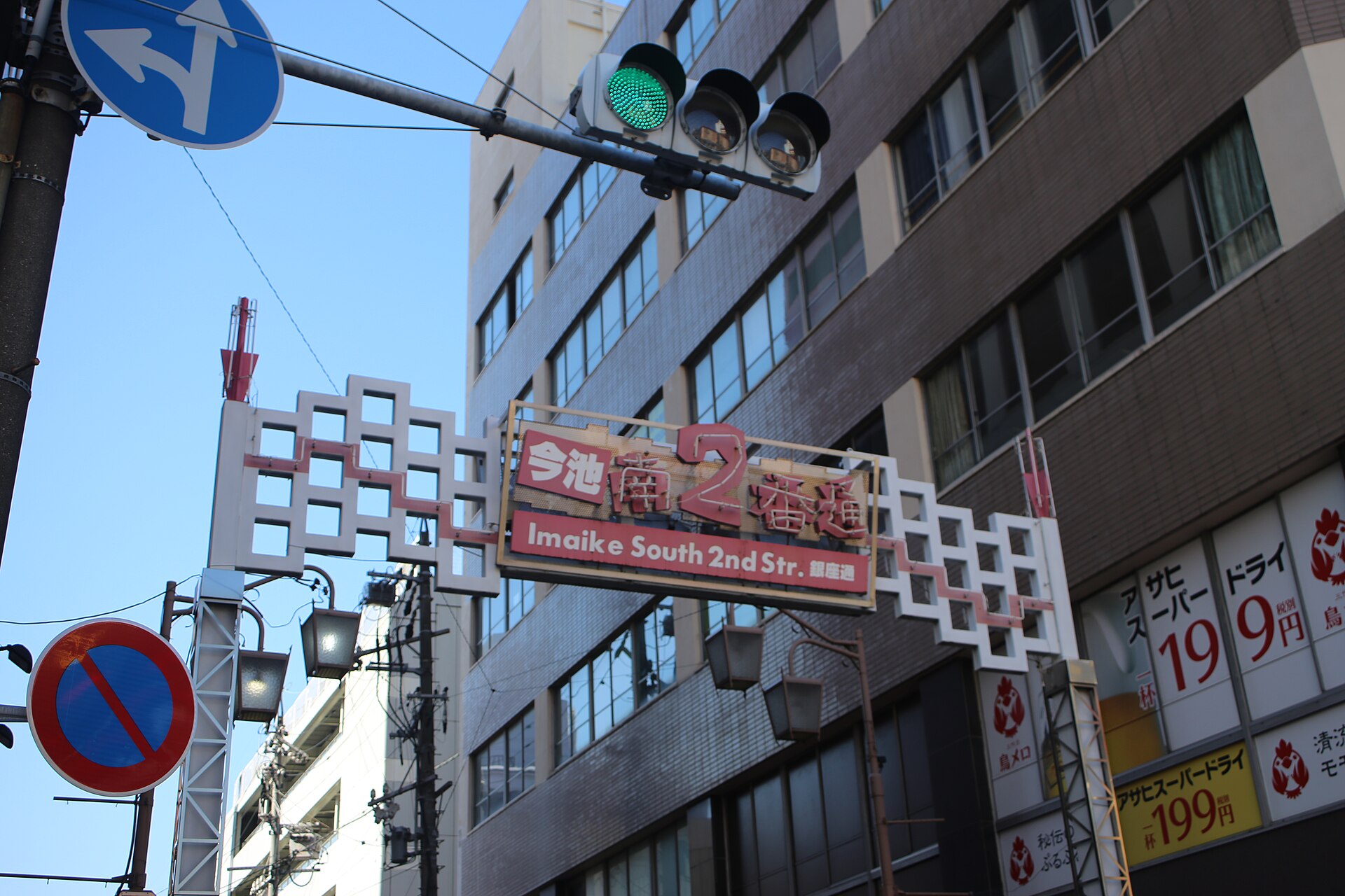 街道上跨着一道白色镂空拱架,红字牌写着「今池南2番通」,拱架上方伸着信号灯与蓝色指路牌