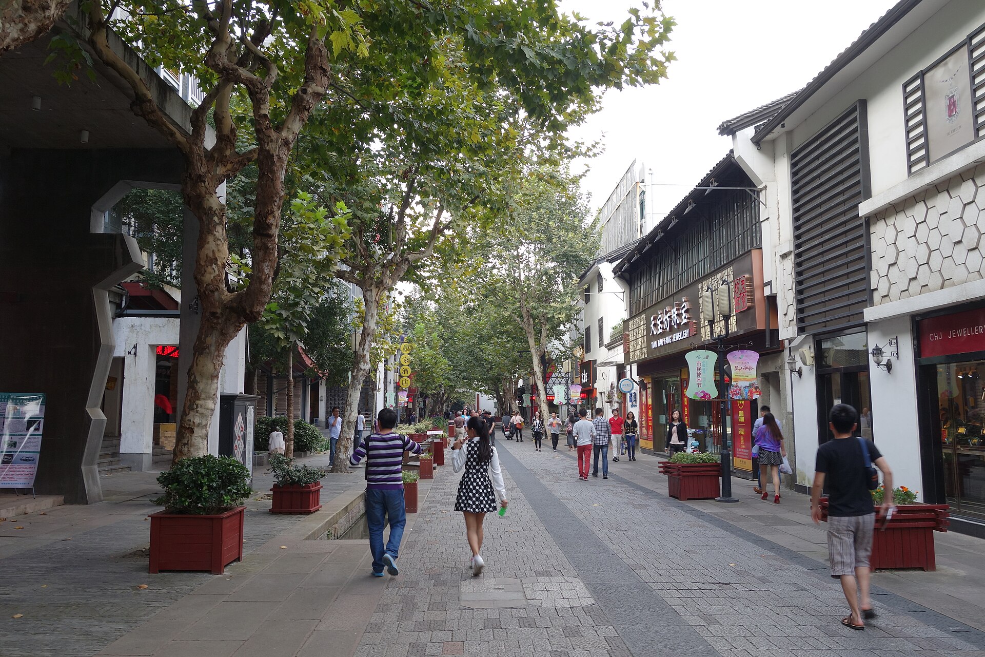 南宋御街的铺装步行街：街道两侧是沿街店铺与成排行道树，行人在石板路面上往来，画面为开阔的商业步行街街景。