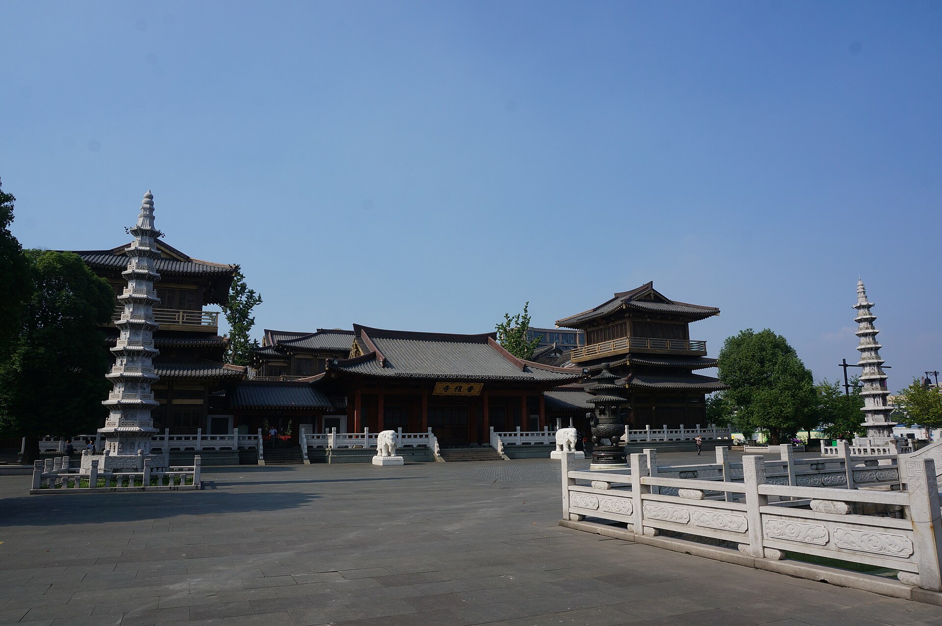 深灰瓦大殿居中，殿前对称摆着石狮与白石栏杆，左侧一座多层石塔、右侧两层重檐楼阁，旁立大型铜香炉