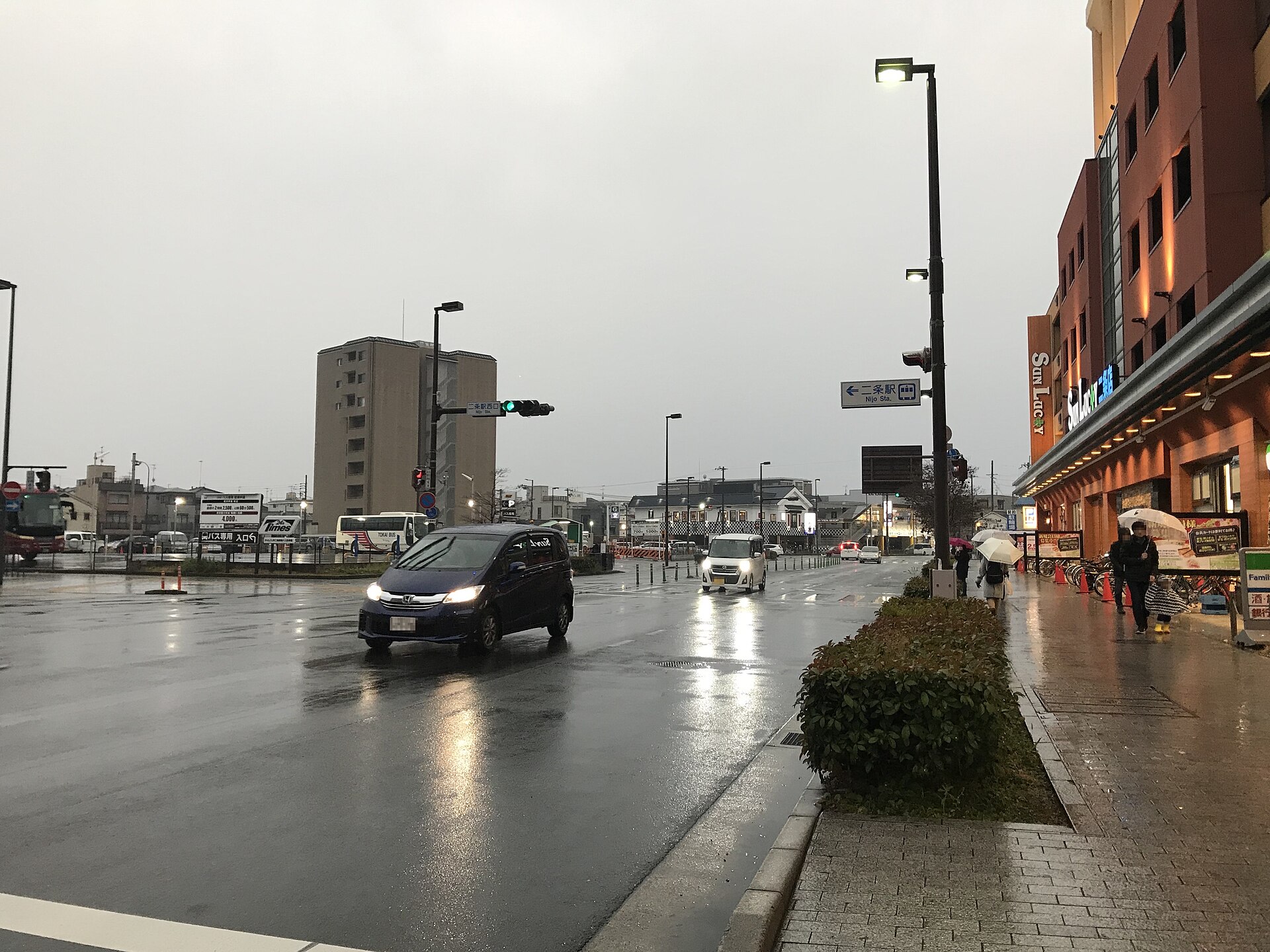 雨天的宽阔路口，一辆深色厢式车驶过湿掉的柏油路面，空中路牌写着「三条駅」「Sanjo Sta.」，两侧是沿街楼与绿篱