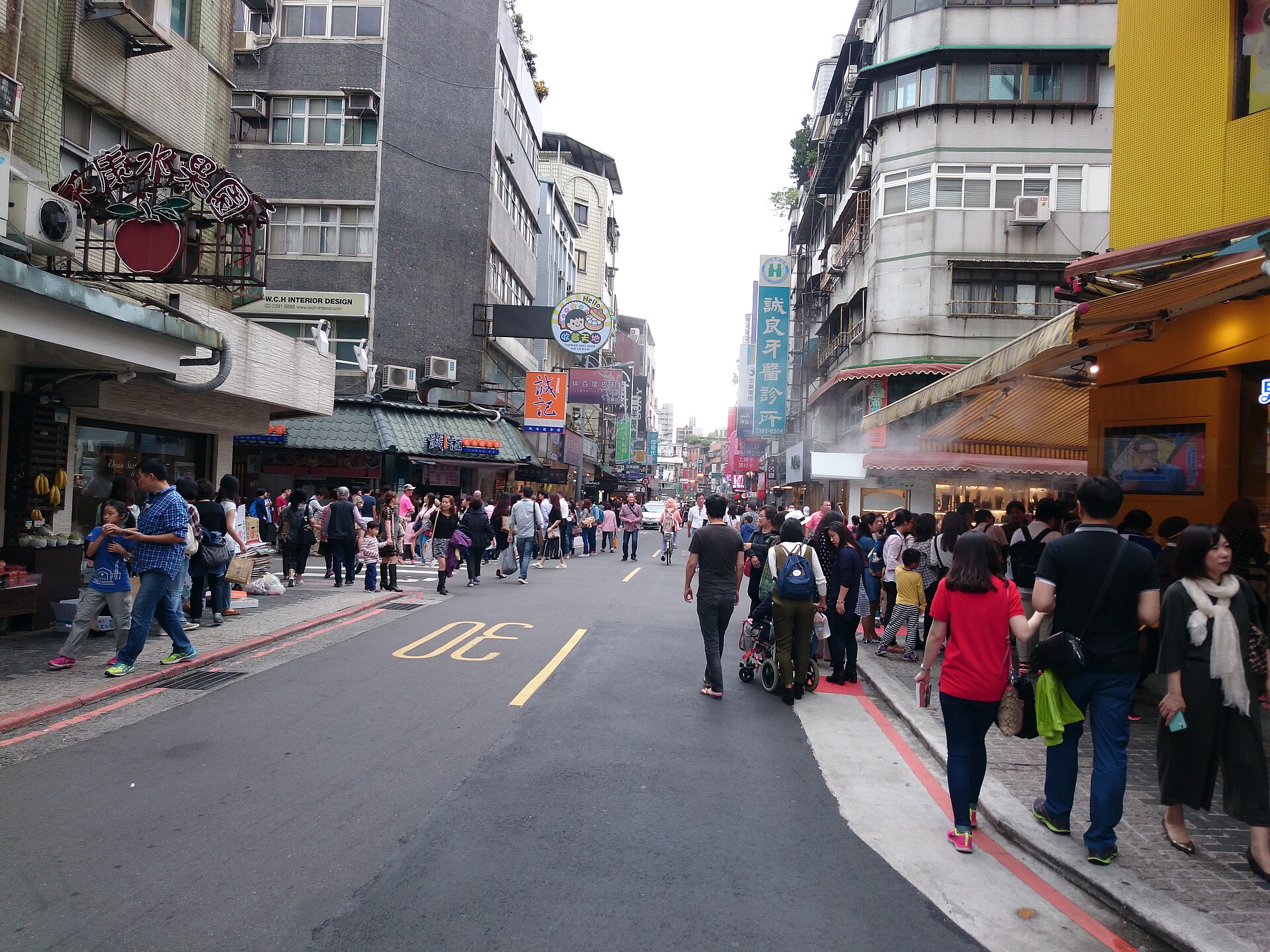 窄街两侧是底层带雨遮的旧楼，路面画着黄色标线并写着「30」，行人提着袋子走，左侧招牌写着「永康水菓園」