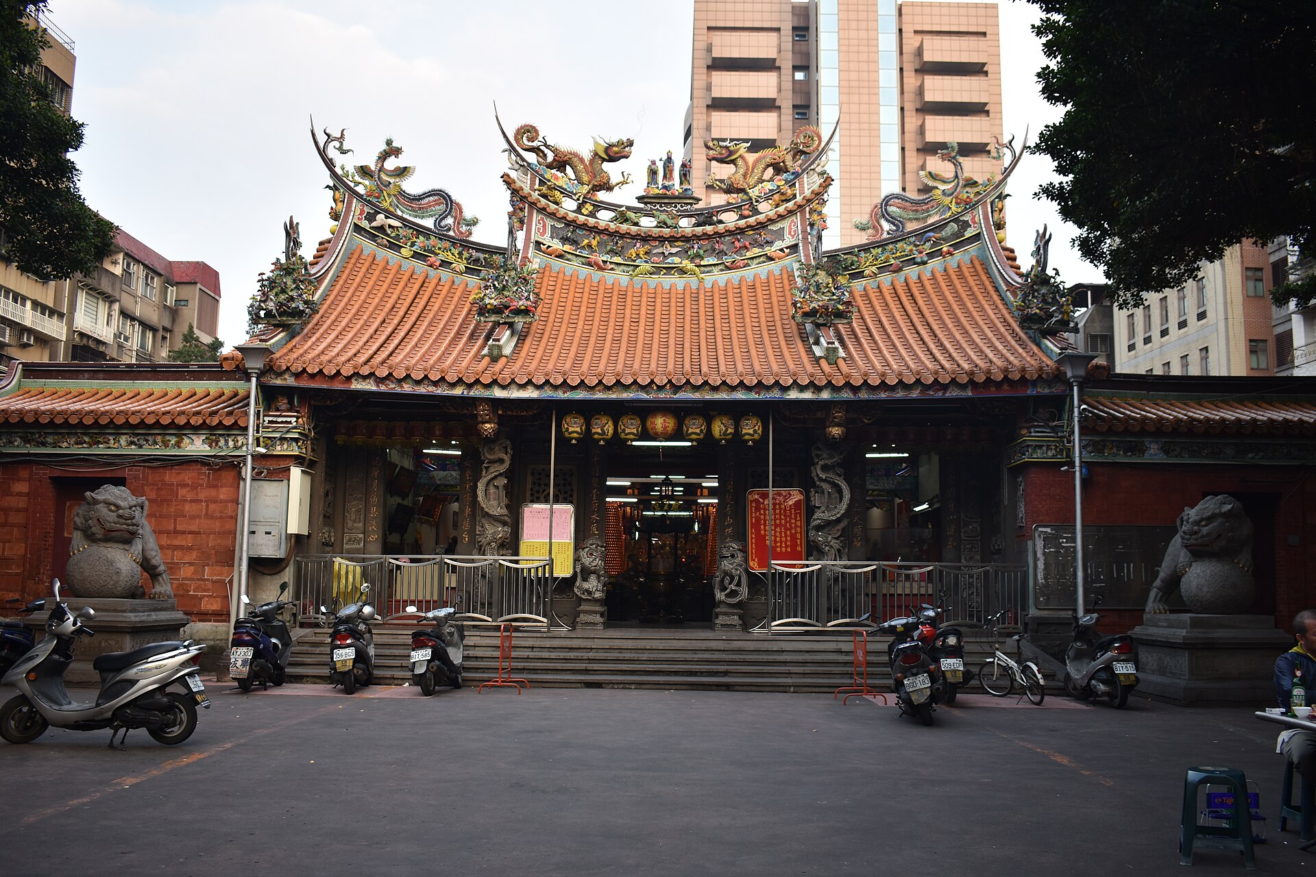 庙宇正面的燕尾脊上立着剪黏龙兽与人物，台阶前守着石狮，庙前柏油广场上停着一排机车与自行车