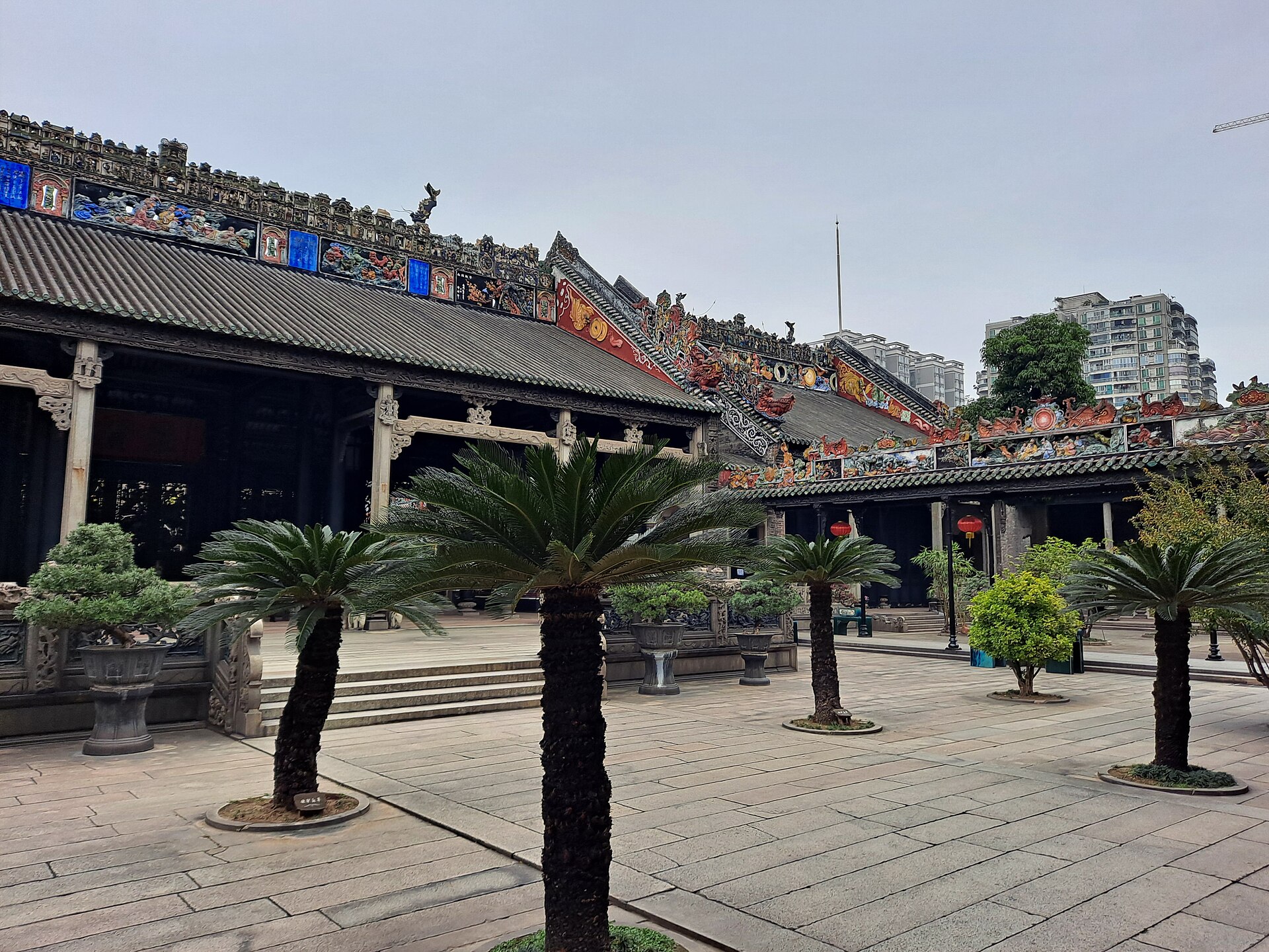祠堂的灰瓦屋脊上密布彩色陶塑与灰塑人物，檐下是深色木构梁架与石柱，庭院石板上摆着多盆苏铁与盆景
