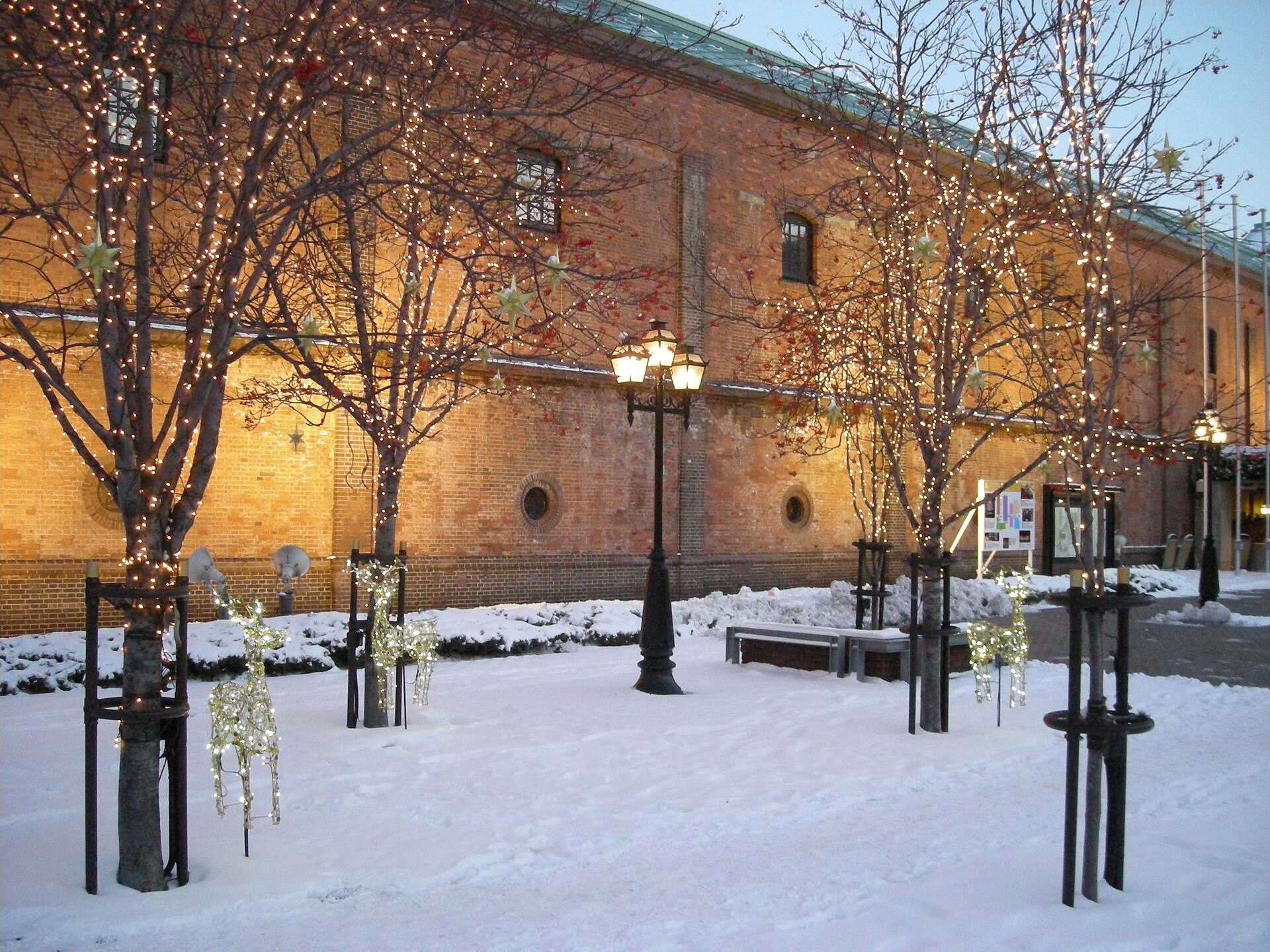 夜里积雪的广场：红砖楼墙上开着拱窗和圆窗洞，檐下射灯照暖砖面，树都缠着灯串