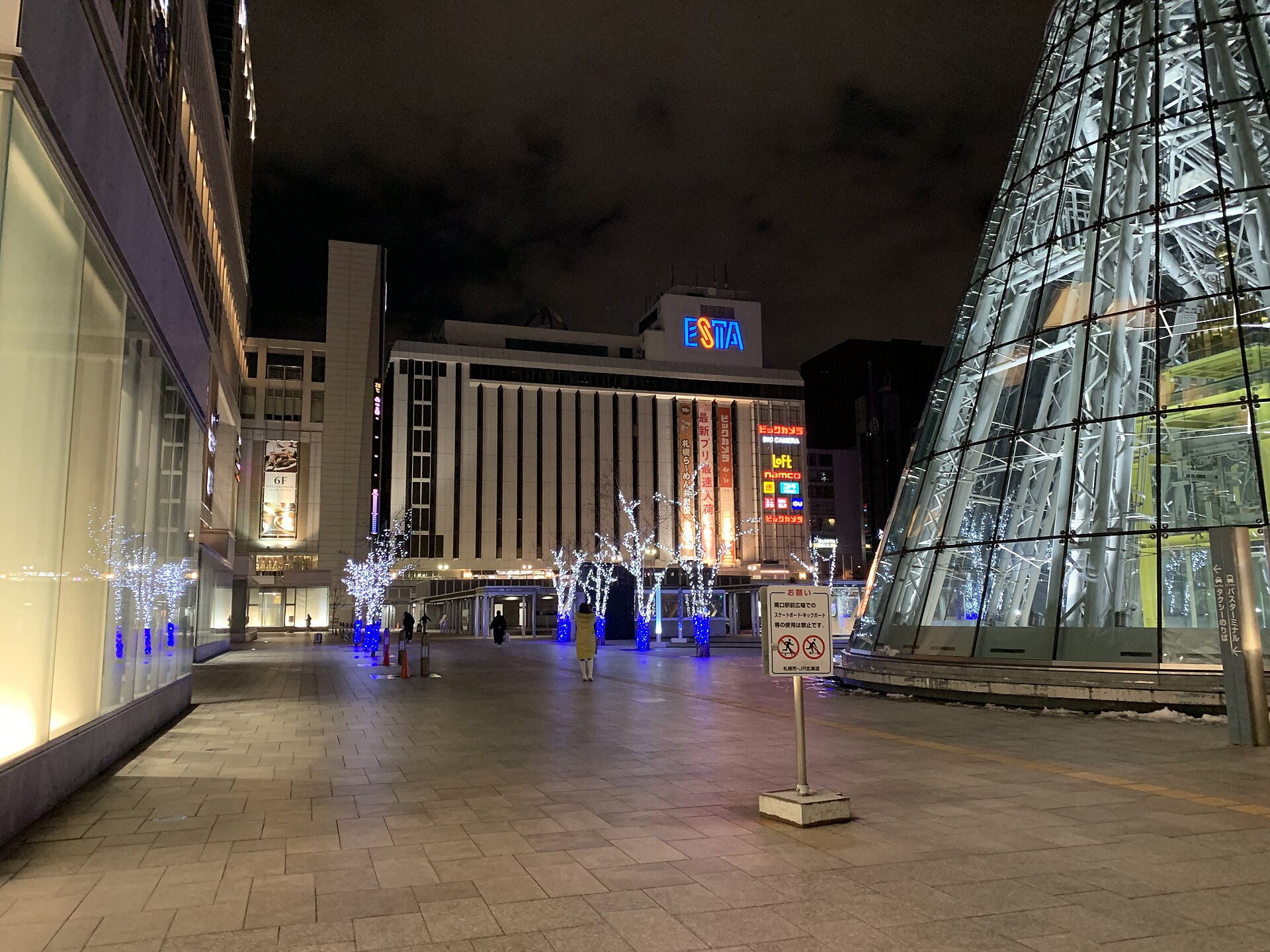 冬夜札幌站南口的站前广场，玻璃锥形出入口泛着灯光，对面商场招牌通明，行道树挂满蓝白灯饰，地面残留薄雪。