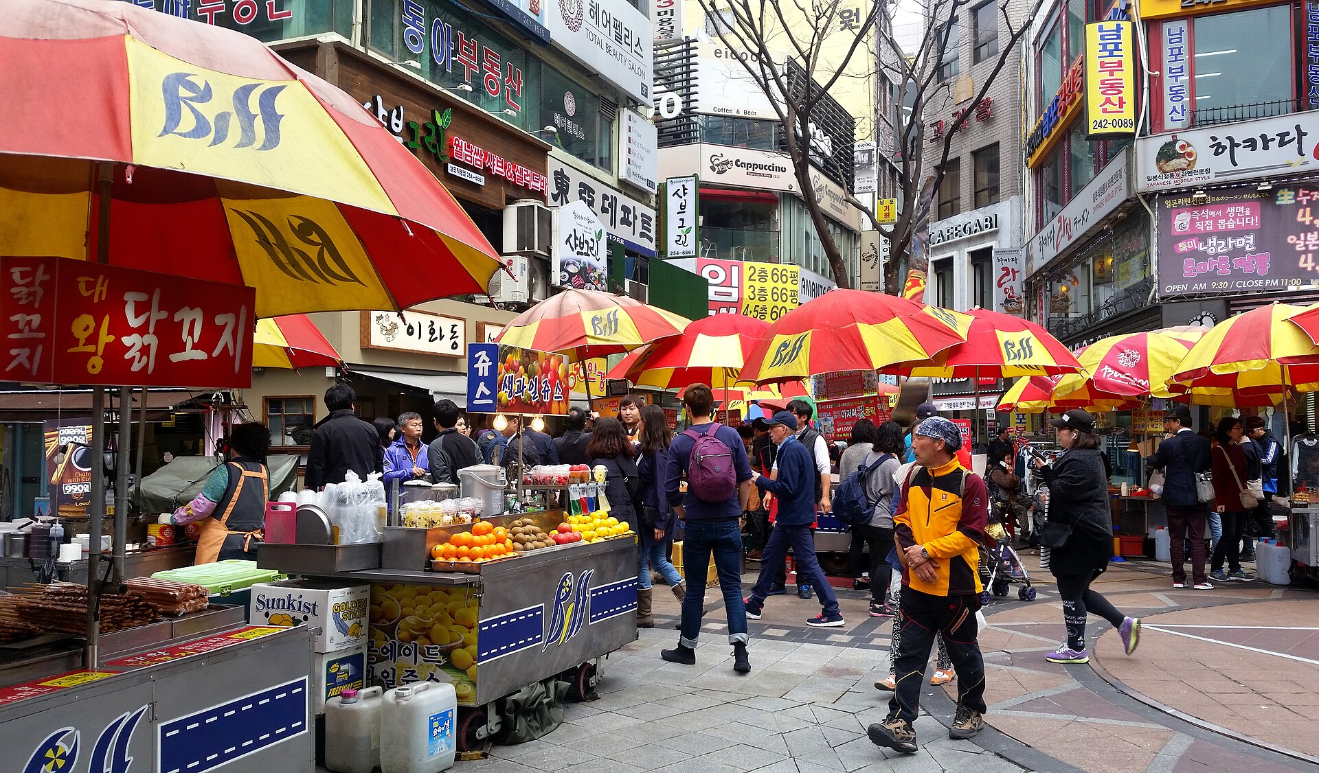 步行街上撑开一长排红黄遮阳伞小吃摊,摊上摆着水果和成摞纸杯,身后商铺挂满韩文招牌(可见「닭꼬치」「닭강정」)