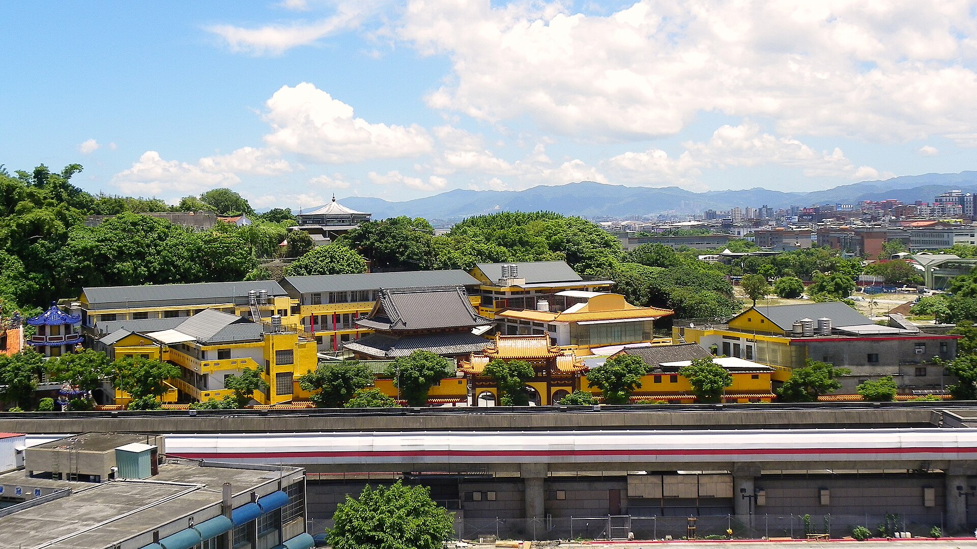 从高处俯看依山而建的黄墙寺院建筑群，左侧山丘上有一座白色圆顶塔，前景一道高架轨道横贯画面