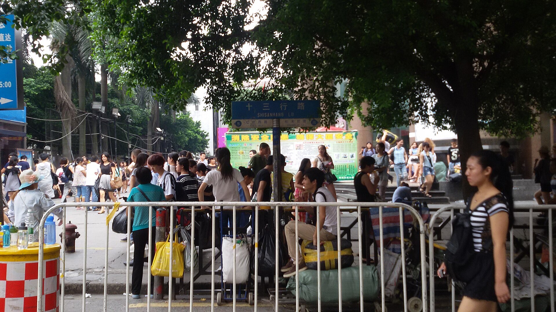 蓝色路牌写着「十三行路」，牌下人行道挤满背着双肩包的人，路边堆着纸箱、编织袋与手推车