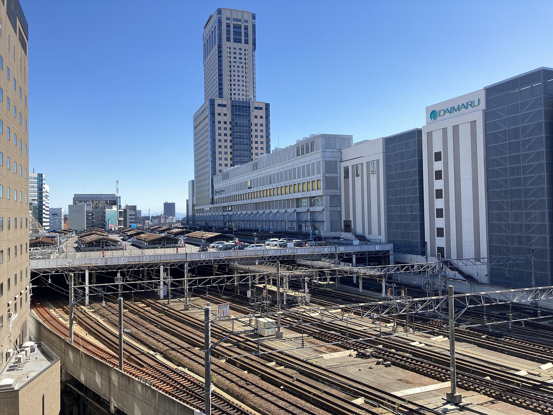 俯瞰站场：成排股道上方横着天桥，桥下挂着 9 到 1 的站台编号，右侧白楼上是「DAIMARU」招牌