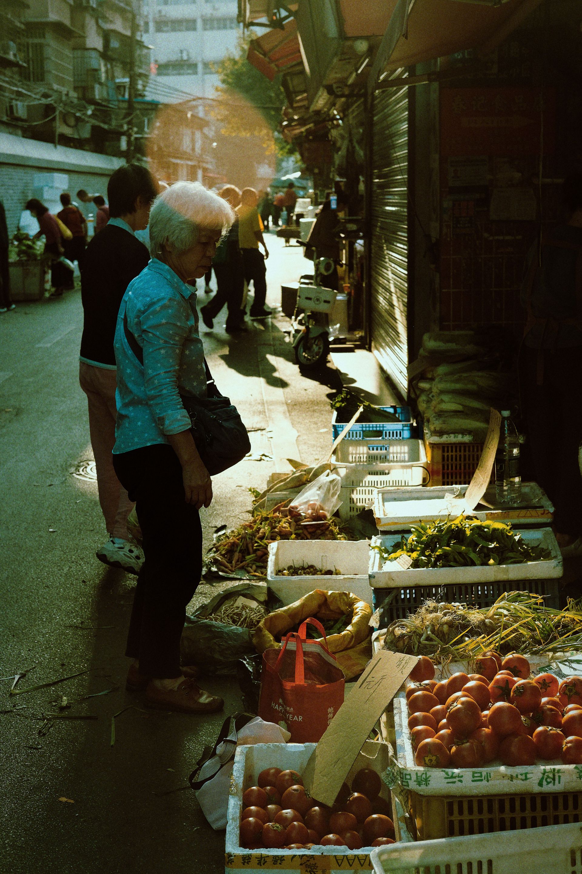 Elderly woman browsing fresh produce at a vibrant street market in Guangzhou, China during twilight.