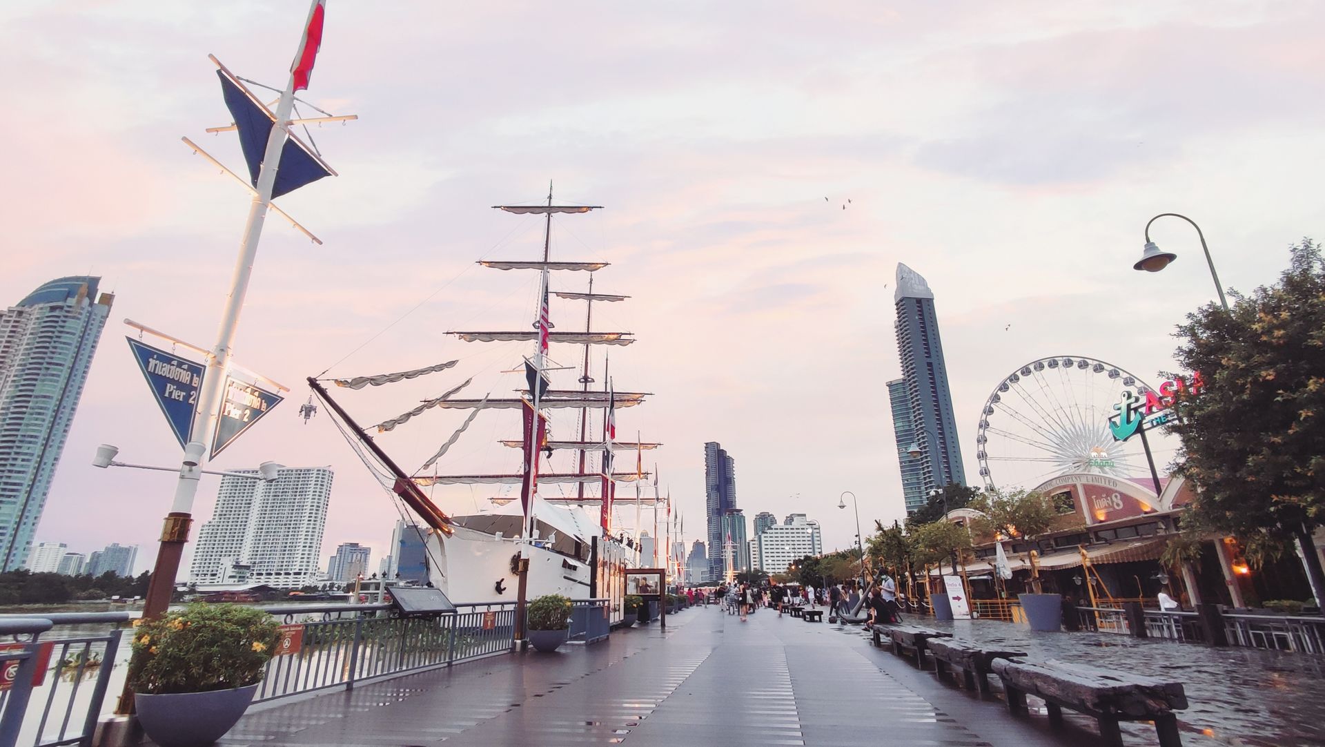 A picturesque view of Bangkok harbor with a tall ship, ferris wheel, and skyline at sunset.