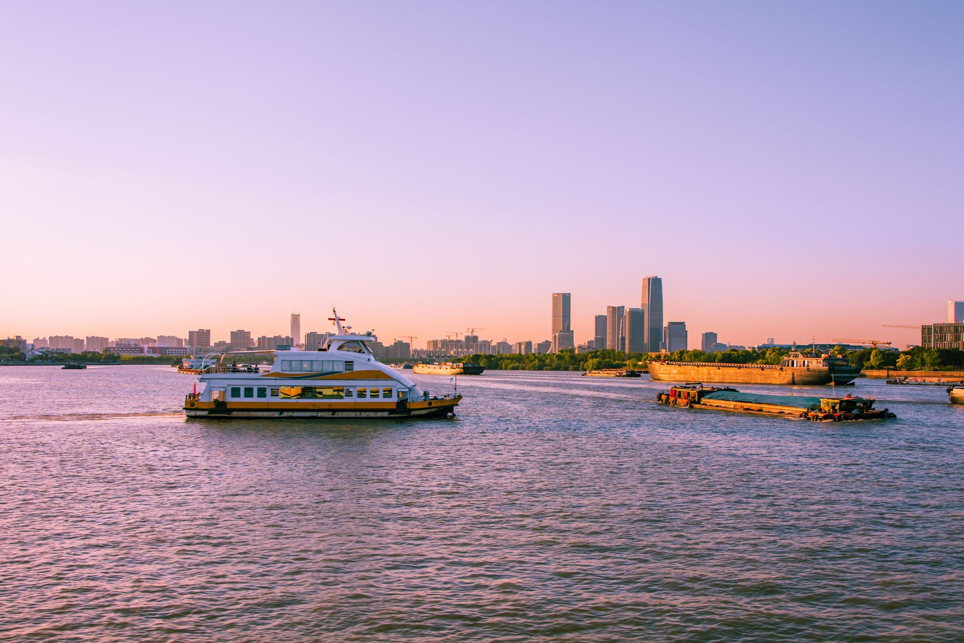 A ferry cruises along a river with a modern city skyline at sunset, reflecting vibrant hues.