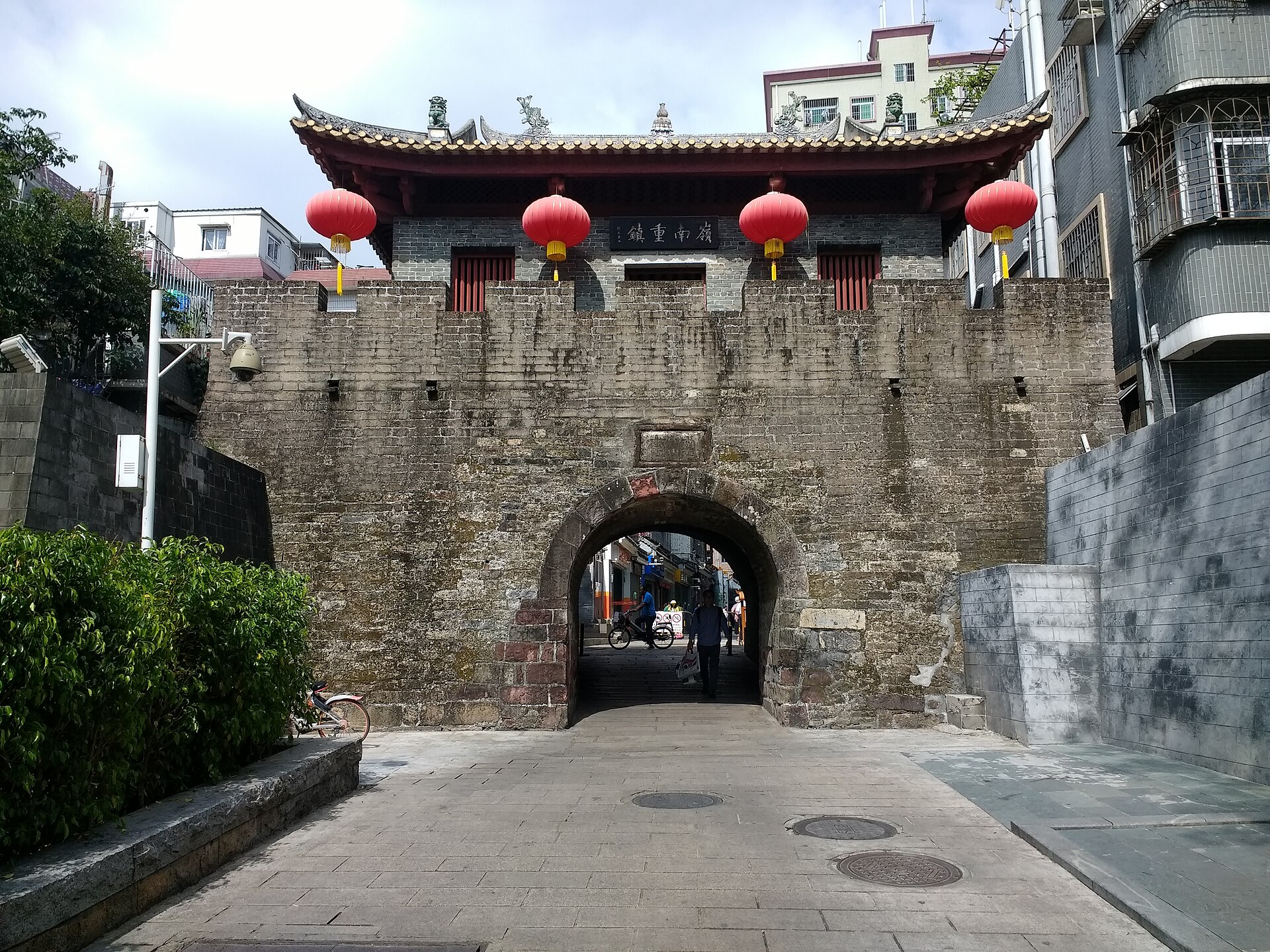 砖石城门开着拱形门洞，檐下横挂匾额「嶺南重鎮」、四只红灯笼分列两侧，城门左右紧贴贴瓷砖的楼房与空调外机